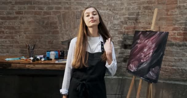 Retrato de talentosa artista femenina bonita con pincel y su lienzo en el fondo. La joven pintora contemporánea mira a la cámara en movimiento con una sonrisa. Chica atractiva en el estudio creativo — Vídeos de Stock