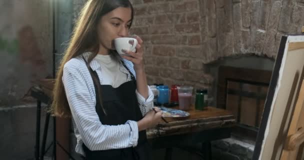 Talentosa artista femenina bonita innovadora dibuja con pincel en el lienzo y beber café. Joven pintora contemporánea creando cuadro abstracto. Muchacha atractiva crea pintura de arte moderno — Vídeos de Stock