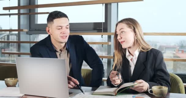 Man and a woman discussing work in the brightly lit modern office. Concerned male and female working with laptop and charts papers at table. Business people discuss new startup project concept — Stock Video