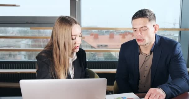 Man and a woman discussing work in the brightly lit modern office. Concerned male and female working with laptop and charts papers at table. Business people discuss project concept. Moving camera — 비디오