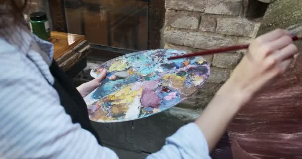 Close-up van de vrouw hand mengen olieverf op houten palet. Aantrekkelijk meisje creëert moderne kunstschilderkunst. close-up vrouwelijke kunstenaar hand met penseel — Stockvideo