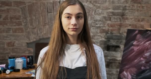 Retrato de talentosa artista femenina innovadora con pincel y su lienzo en el fondo. La joven pintora contemporánea mira a la cámara con una sonrisa. Chica atractiva en el estudio creativo — Vídeos de Stock