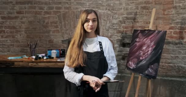 Portrait of talented pretty female artist with brush and her canvas on background. Contemporary young woman painter looks at the moving camera with a smile. Attractive girl in creative studio — 비디오