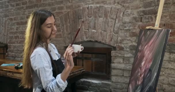 Talento innovativo graziosa artista femminile disegna con pennello sulla tela e bere caffè. Giovane pittrice contemporanea che crea quadri astratti. Attraente ragazza crea arte moderna pittura — Video Stock