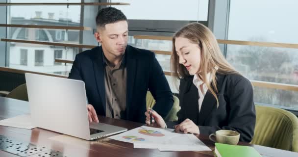 Man and a woman discussing work in the brightly lit modern office. Concerned male and female working with laptop and charts papers at table. Business people discuss new startup project concept — Stock Video