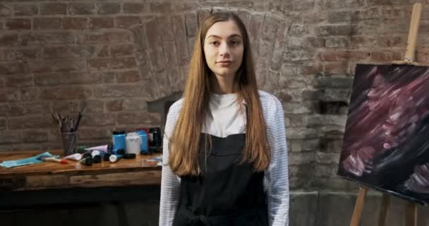 Retrato de talentosa artista femenina innovadora con pincel y su lienzo en el fondo. La joven pintora contemporánea mira a la cámara con una sonrisa. Chica atractiva en el estudio creativo — Vídeos de Stock