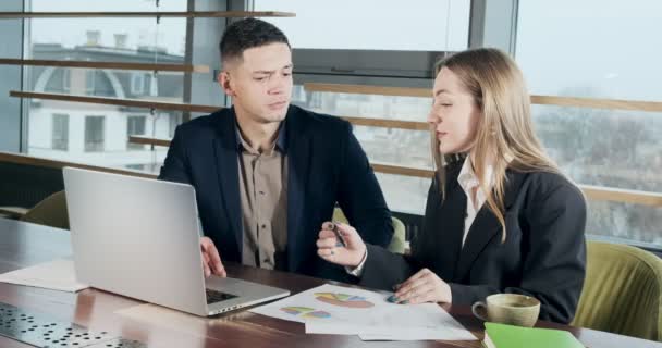 Man och kvinna diskuterar arbete på det starkt upplysta moderna kontoret. Berörda manliga och kvinnliga arbetar med bärbar dator och diagram papper vid bordet. Affärsmän diskuterar nytt koncept för uppstartsprojekt — Stockvideo