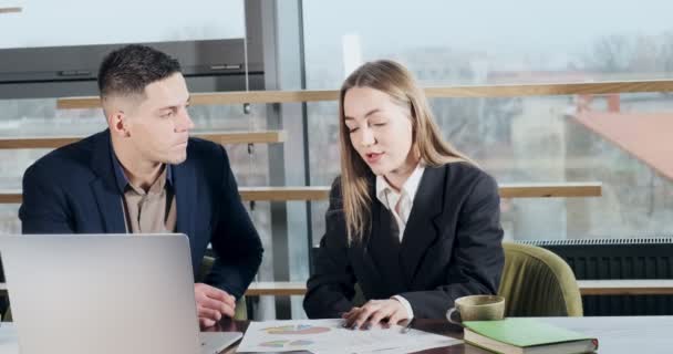 Hombre y una mujer discutiendo el trabajo en la oficina moderna brillantemente iluminada. Preocupado hombre y mujer que trabajan con el ordenador portátil y cartas papeles en la mesa. Gente de negocios discuten nuevo concepto de proyecto de startup — Vídeos de Stock