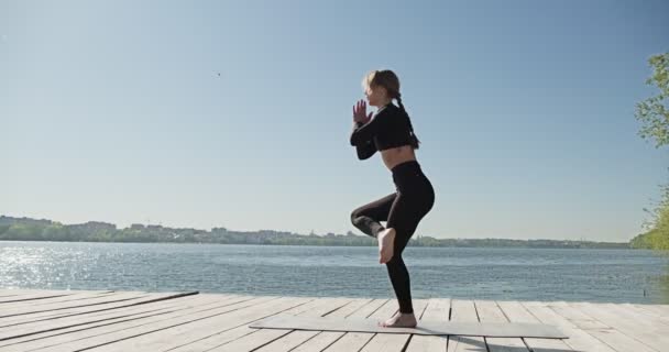 Giovane donna bionda che pratica yoga sul letto di legno al lago. Singolo sport sano allenamento sulla natura al tempo soleggiato — Video Stock