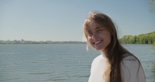 Πορτρέτο της νεαρής ελκυστική γυναίκα που χαμογελά και κοιτάζοντας την κάμερα. Χαρούμενα όμορφη γυναίκα κοντά στη λίμνη σε ηλιόλουστο καιρό. Κλείσε. — Αρχείο Βίντεο
