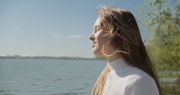 Gülümseyen ve kameraya bakan çekici genç bir kadının portresi. Güneşli havalarda göl kenarında neşeli, güzel bir kadın. Kapat. — Stok video