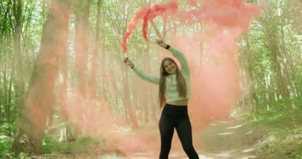 Menina bonita em uma floresta verde gira ao redor enquanto segurando duas bombas de fumaça vermelha. Movimento lento — Vídeo de Stock