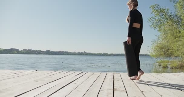 Ung blond kvinna praktiserar yoga på träkajen vid sjön. Singel sport hälsosam träning på naturen vid soligt väder — Stockvideo