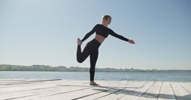 Genç sarışın kadın göldeki ahşap ranzada yoga yapıyor. Güneşli havalarda tek spor doğa üzerine sağlıklı eğitim