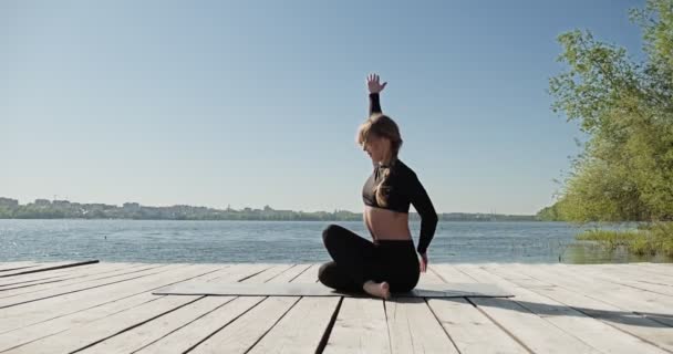 Jonge blonde vrouw die yoga beoefent op de houten ligplaats aan het meer. Single sport gezonde training op de natuur bij zonnig weer — Stockvideo