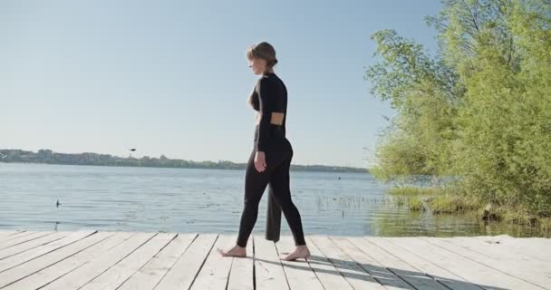 年轻的金发女人在湖边的木床上练习瑜伽。在阳光普照的天气下进行关于自然的单项运动健康训练 — 图库视频影像