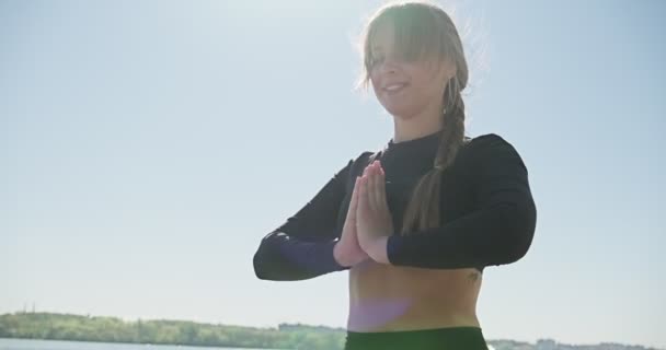 Mladá blondýnka cvičí jógu na dřevěném lůžku u jezera. Jednotný sport zdravý trénink na přírodě za slunečného počasí — Stock video