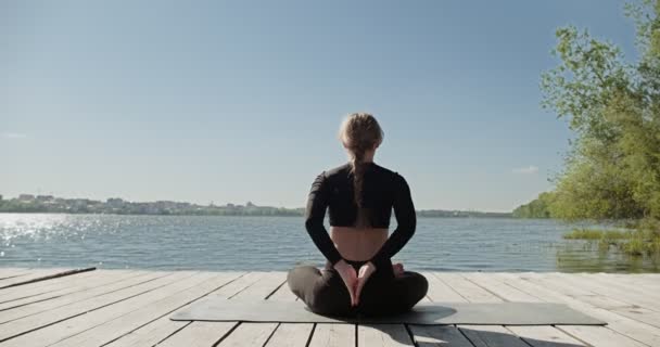 Jonge blonde vrouw die yoga beoefent op de houten ligplaats aan het meer. Single sport gezonde training op de natuur bij zonnig weer — Stockvideo