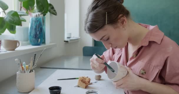 Γυναίκα αγγειοπλάστη κάθεται και ανακατεύει χρώμα με ένα πινέλο ένα φλιτζάνι στο τραπέζι. Γυναίκα που φτιάχνει κεραμικά αντικείμενα. Εργασίες αγγειοπλαστικής, χειροποίητες και δημιουργικές δεξιότητες — Αρχείο Βίντεο