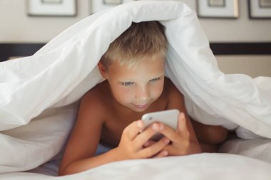Güzel bir beyaz çocuk bir battaniyenin altında yatakta yatıyor ve bir gadget'ı elinde tutar.