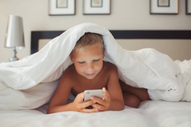Güzel bir beyaz çocuk bir battaniyenin altında yatakta yatıyor ve bir gadget'ı elinde tutar.