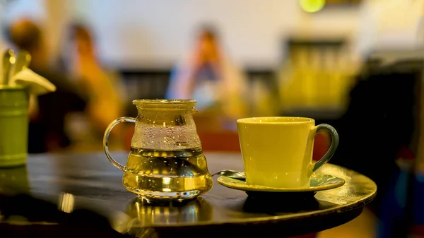 Tasse Kaffee oder Tee, Glas Teekanne mit heißem Wasser, morgens. Bild mit verschwommenen Cafébesuchern in leiser Tonart, zur Hintergrundnutzung — Stockfoto