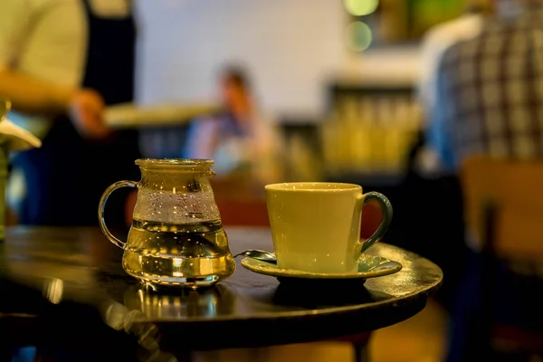 Φλιτζάνι καφέ ή ΤΕΕ, γυάλινη τσαγιέρα του ζεστού νερού, πρωί. Εικόνα με θολή καφέ επισκέπτες και σερβιτόρος σε μια χαμηλών τόνων, για το φόντο. Έννοια του τρόπου ζωής — Φωτογραφία Αρχείου