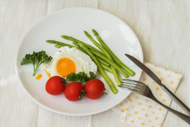 Lezzetli sağlıklı gurme kahvaltı, geleneksel ev yapımı kızarmış yumurta ve taze kuşkonmaz peçete, çatal ve bıçak ile beyaz tabakta domates ile servis