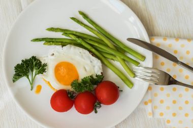 Taze kuşkonmaz ile yakın çekim kızarmış yumurta, peçete, çatal ve bıçak, sabah yemek ile beyaz tabakta domates. Üstten Görünüm