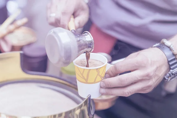 Gros plan des mains de l'homme qui fait du café turc traditionnel en turque de cuivre sur sable chaud, se déverse dans une tasse en papier jetable. Concept de préparation du café, fond tonned — Photo