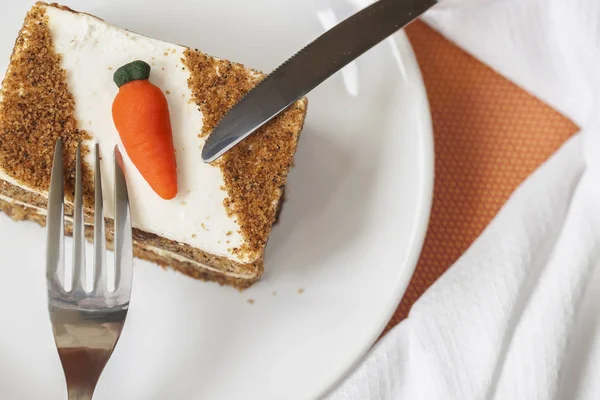 Homemade carrot cake with carrot decorations on white plate, fork, knife, napkin, top view.