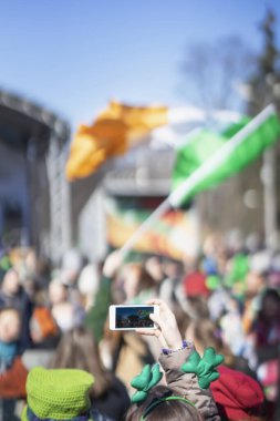 Female hands with mobile phone, making photo of traditional national Irish holiday party, carnival of St. Patricks Day on a smartphone, selective focus. clipart