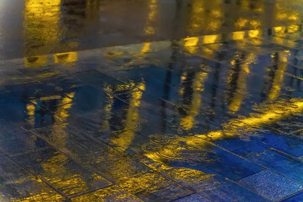 Blurred reflection of a bright building in a puddle on the asphalt. Rain in the city at night. Abstract vivid colorful background. Selective focus — ストック写真