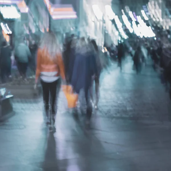 Abstract meisje in een fel oranje jasje terug naar ons, donkere wazige achtergrond, stad straat in de avond, voetgangersstraat, ongeïdentificeerde mensen, ongeconcentreerde beweging — Stockfoto