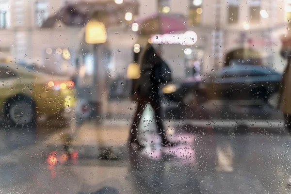 Blurry reflection silhouettes of the people in walking on a rain under umbrellas and bokeh city lights, night. View through glass window with rain drops — Stock Photo, Image