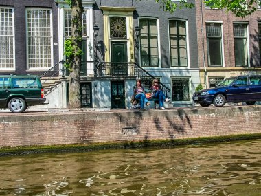 Amsterdam, Hollanda - 5 Mayıs 2007: Güzel evlerin arka planındaki kanal bankasında iki kız ve iki araba