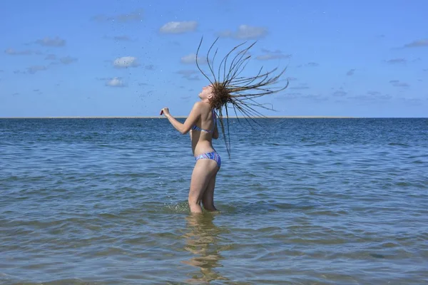 Uma menina está de pé no mar e jogando seu longo cabelo molhado para trás, água espirrando no ar — Fotografia de Stock