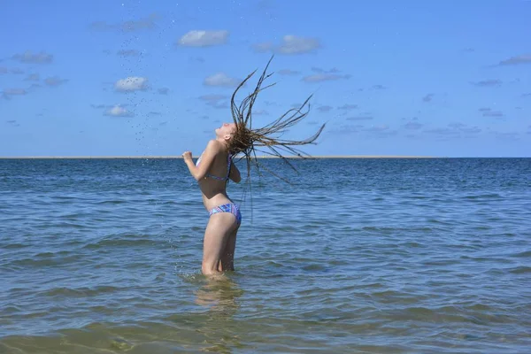 Junges Mädchen steht im Meer und wirft ihre langen nassen Haare zurück, Wasser spritzt in die Luft — Stockfoto