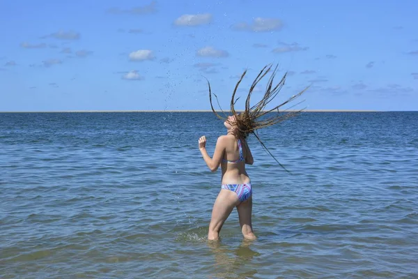 Uma menina está de pé no mar e jogando seu longo cabelo molhado para trás, água espirrando no ar — Fotografia de Stock