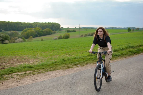 A young woman , sits on a bike and drives on a lonely country road in green nature with blue sky