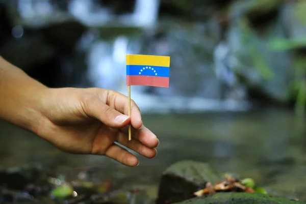 La pobre Venezuela en manos de gente poderosa. Un hombre torcido sostiene la bandera de Venezuela cerca del arroyo. Concepto de dominación y criminalidad. Prueba de depravación y avaricia. Problemas con los drogs . — Foto de Stock