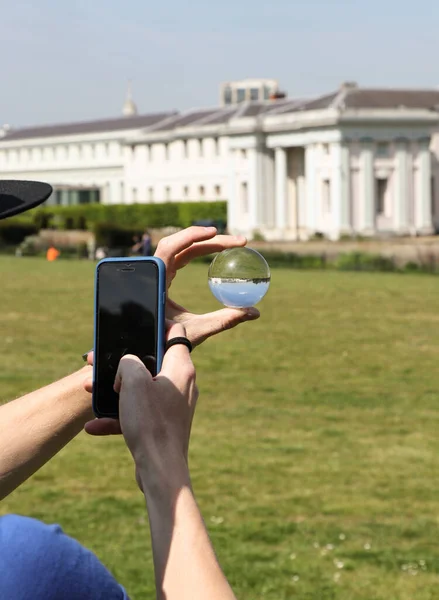 Young guy take a picture with his phone across lens ball. Learn to take pictures with glass ball in greenwich park in London, Great Britain. City landscape with phone and crystal ball.