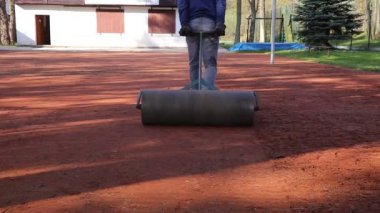 Genç yaz işçisi turnuvaya ve lig voleybol maçlarına hazırlanıyordu. İşçi arkasından ağır bir silindir çeker ve kilden boğulur. Bahçıvanlık. Sarayın yüzeyini ayarlayın.