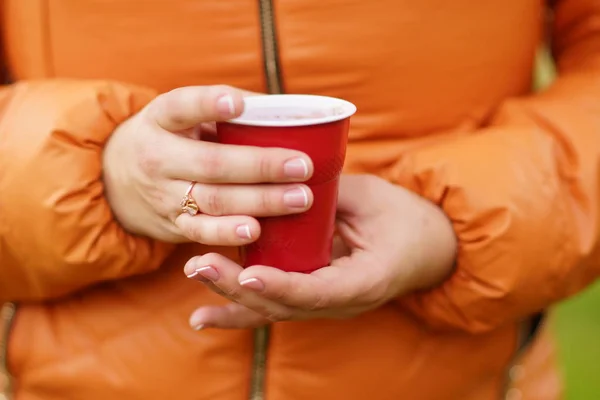 Руки девушки в оранжевой куртке держат красную одноразовую чашку с кофе, чаем, латте, капучино — стоковое фото