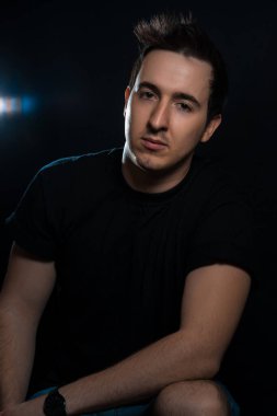 Kollarını kavuşturmuş yakışıklı genç bir adam siyah bir arka planda oturur ve kameraya bakar.