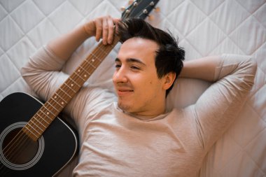 Yumuşak odaklanma. Hafif kazaklı ve kot pantolonlu genç bir adam sabah evde yatakta akustik gitar çalar.