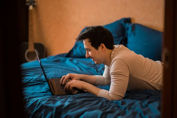 Man using laptop in a hotel room or home bedroom. The guy is lying on the bed and typing from a laptop. Man in the morning with a modern device, reading emails and sending messages. Distance learning during the Covid-19 pandemic. freelasner, distant