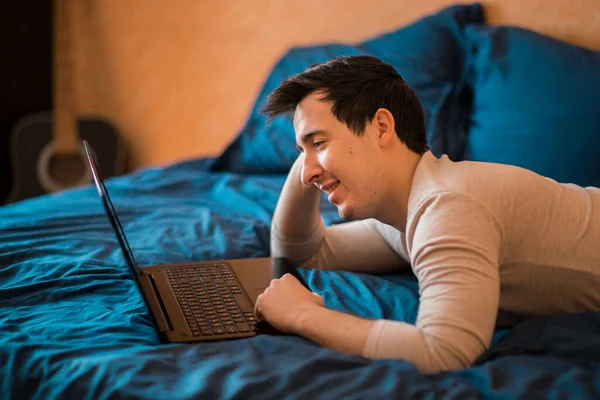 Man using laptop in a hotel room or home bedroom. The guy is lying on the bed and typing from a laptop. Man in the morning with a modern device, reading emails and sending messages. Distance learning during the Covid-19 pandemic. freelasner, distant