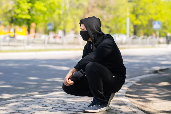 Caucasian teenager boy in facial medical mask sitting near the edge of the roadway. teen in a medical mask on the street