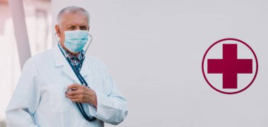 Beyaz önlüklü ve yüzünde tıbbi maske olan gri saçlı yaşlı bir erkek doktor hastaların akciğerlerini ve kalplerini incelemek için steteskop kullandığını gösteriyor..
